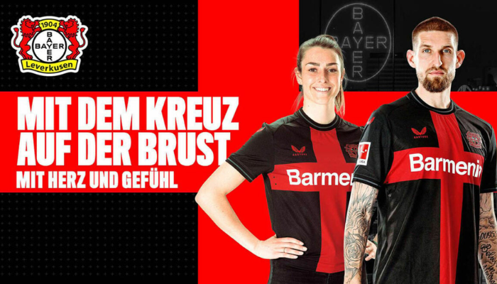 Camisa titular do Bayer Leverkusen Castore 2023-2024. A camisa é predominantemente preta com detalhes em vermelho e branco. No centro do peito, há uma cruz vermelha, símbolo da Bayer. A gola redonda e os punhos são pretos com finas linhas cinza. Na nuca, está escrito "Werkself" em vermelho. 